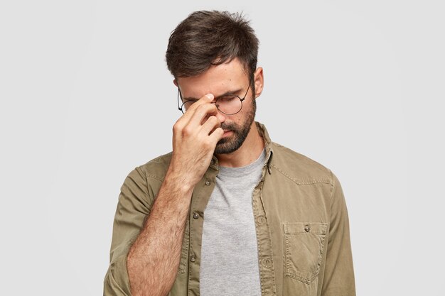 Sad unhappy beared man keeps hand on nose, head down, tries to concentrated, has overworked expression