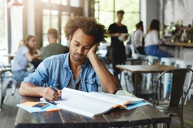 Sad unhappy dark-skinned bearded student feeling frustrated while preparing for lessons at college, writing down in his copybook with pen, leaning on elbow and looking at notes with upset expression