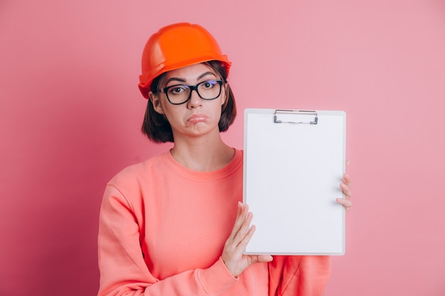 슬픈 불행 실망 된 여자 작업자 작성기 분홍색 배경에 흰색 사인 보드를 빈 개최. 건물 헬멧.