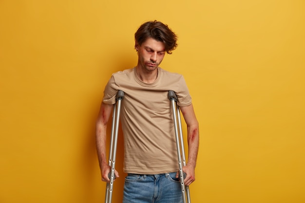 Sad unhappy man looks down, has serious injury after falling down from height, tired of long recovery period, tries to walk with crutches, poses against yellow wall. Handicapped disabled male