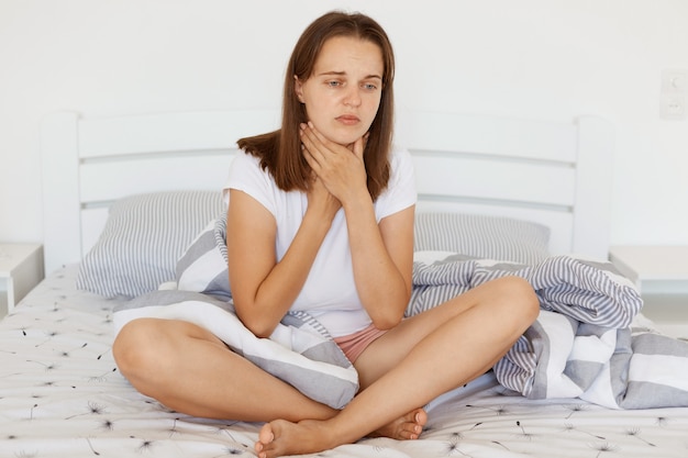 Sad unhappy sick woman with dark hair wearing white casual t shirt sitiing on bed with crossed legs, touching her neck, suffering from sore throat, frowning face.