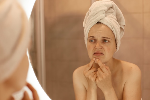 Sad unhappy young adult woman wearing bath towel squeezing acne on chin, mirror reflection of female with pimples on her face, skin problems, skin care.