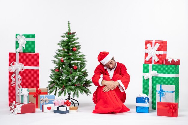 Грустный несчастный молодой человек в костюме Санта-Клауса с подарками и украшенной елкой сидит на земле на белом фоне