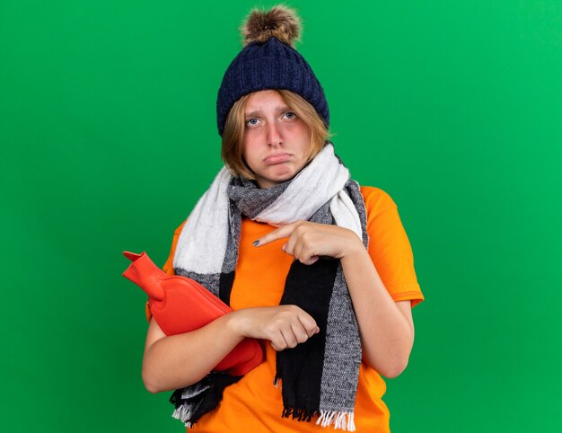 Sad unhealthy young woman in orange t-shirt with hat and warm scarf around neck feeling terrible holding hot-water bottle pointing with finger at it suffering from cold standing over green wall