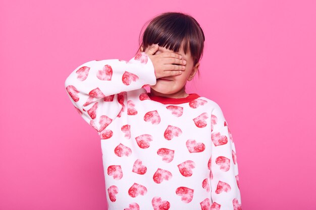 슬픈 화가 어린 소녀는 나쁜 소식, 울고, 손바닥으로 눈을 가리고, 분홍색 벽 위에 고립 된 하트 프린트가있는 흰색 스웨터를 입고 있습니다.