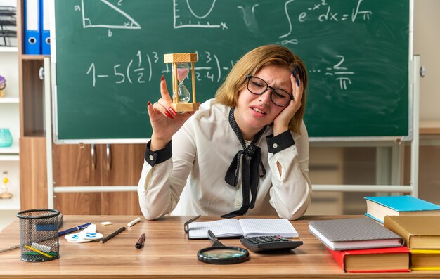 sad with closed eyes young female teacher sits at table with school supplies holding hourglass in classroom