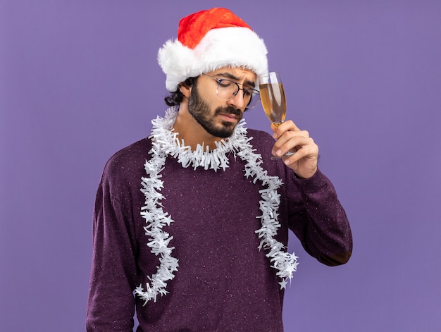 Sad with closed eyes young handsome guy wearing christmas hat with garland on neck holding glass of champagne around forehead isolated on blue background