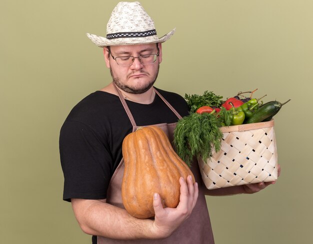 Sad with closed eyes young male gardener wearing gardening hat holding vegetable basket with pumpkin isolated on olive green wall