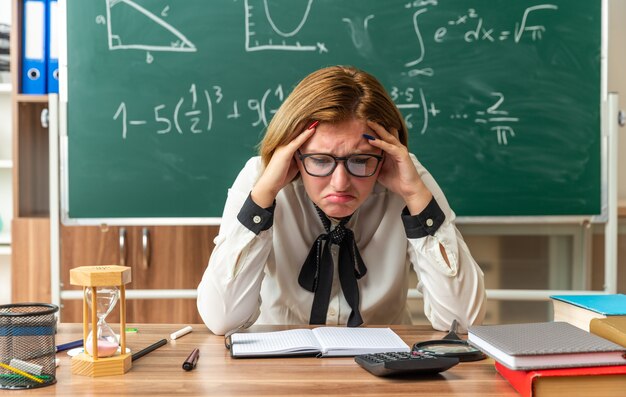 Sad with lowered head young female teacher wearing glasses sits at table with school tools putting hands on head in classroom