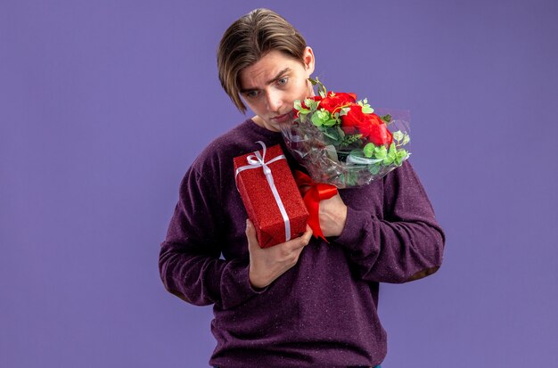 Sad with lowered head young guy on valentines day holding gift box with bouquet isolated on blue background