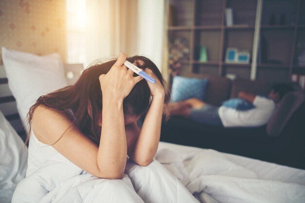 Sad woman complaining holding a pregnancy test sitting on bed