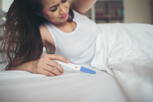 Sad woman complaining holding a pregnancy test sitting on bed