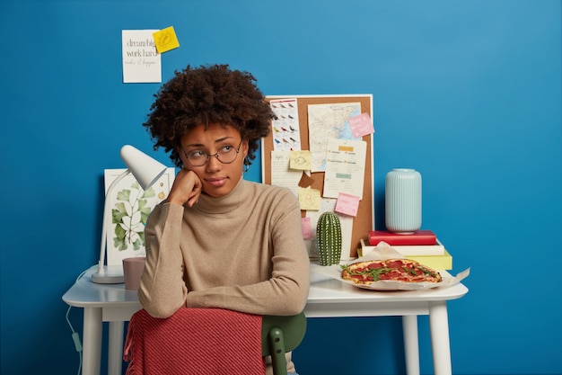 Sad woman has Afro haircut sits at chair, wears round spectacles and beige jumper, sits in coworking space, table with notes desk lamp and delicious pizza behind.