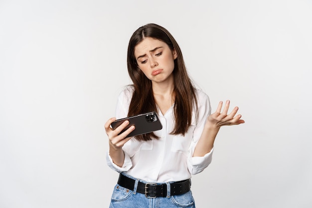 Грустная женщина проигрывает в мобильной видеоигре, выглядит расстроенной и разочарованной на смартфоне, дуется, стоит на белом фоне