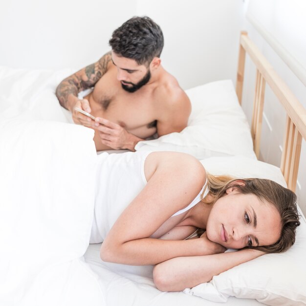 Sad woman lying on bed in front of her husband using mobile phone