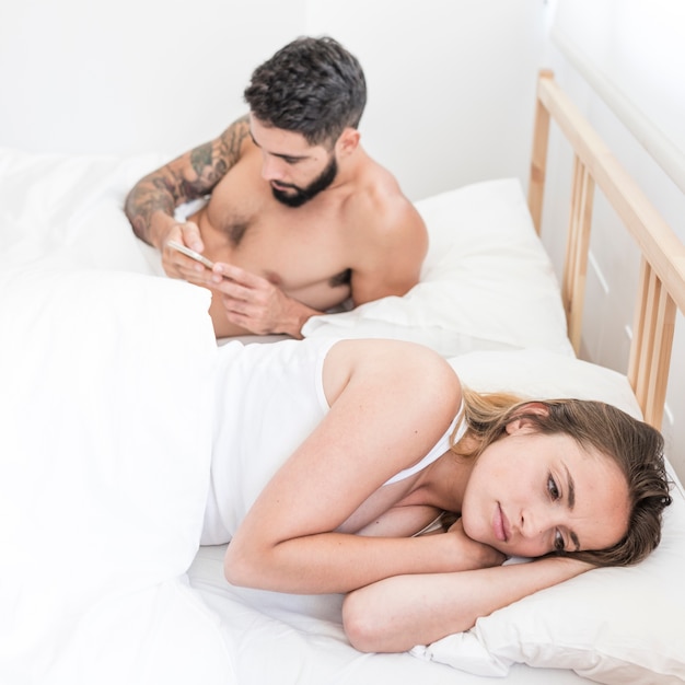 Грустная женщина, лежащая на кровати перед мужем, используя мобильный телефон