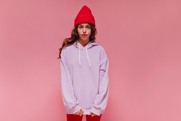 Free photo sad woman in red hat cycling shorts and purple hoodie poses on pink background brunette curly girl looks disappointed and dissatisfied on isolated