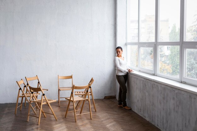 Sad woman next to windowsill at a group therapy session with empty chairs