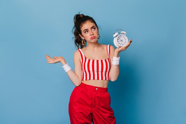 Sad woman with ponytail looks up in dismay, holding white alarm clock in her hands