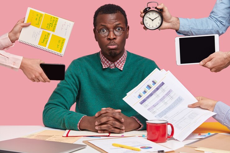 Free Photo sad young african american man dressed in green sweater, sits at desktop, hands with papers, alarm clock, touchpad, notepad with stickes