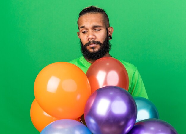 Sad young afro-american guy wearing green t-shirt standing behind balloons isolated on green wall