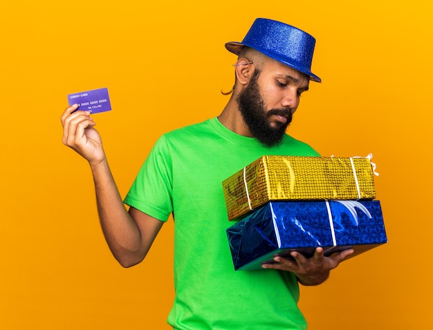 Sad young afro-american guy wearing party hat holding gift boxes with credit card 