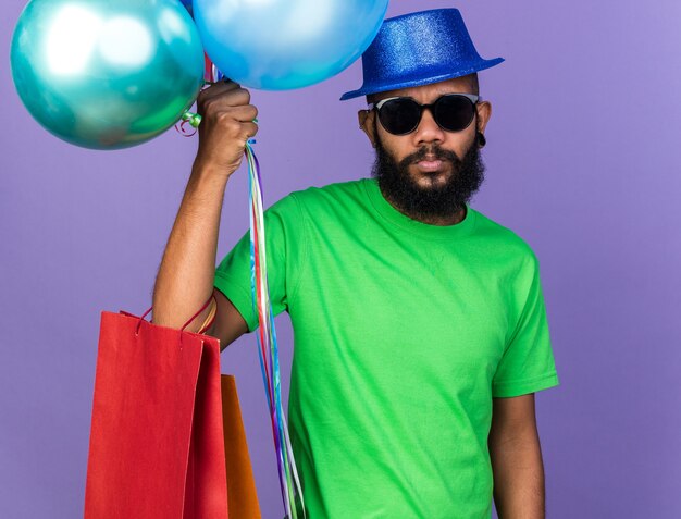 Sad young afro-american guy wearing party hat with glasses holding balloons with gift bag isolated on blue wall