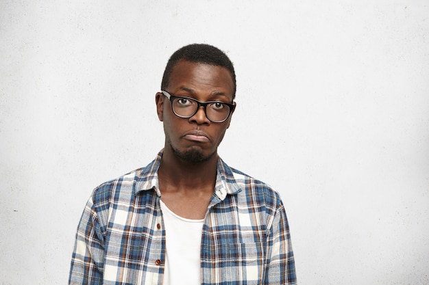 Sad young Afro-American man in trendy shirt and eyeglasses screwing up lips while feeling disappointed and miserable with about his life, standing isolated