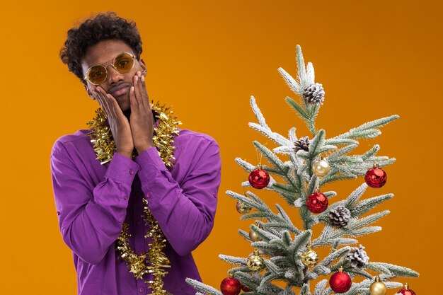 Sad young afro-american man wearing glasses with tinsel garland around neck standing near decorated christmas tree on orange background