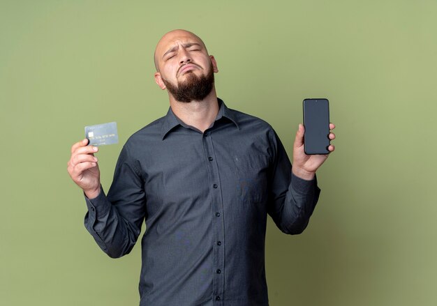 Sad young bald call center man holding mobile phone and credit card with closed eyes isolated on olive green wall