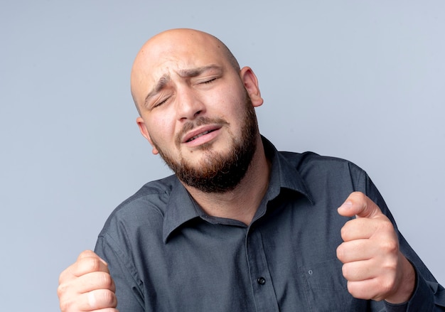 Sad young bald call center man with clenched fists and closed eyes isolated on white wall