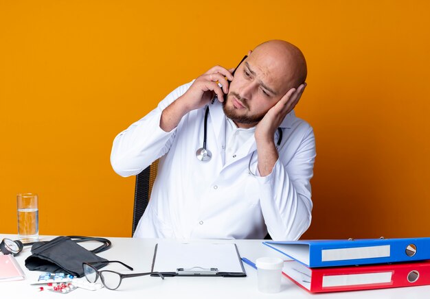 Sad young bald male doctor wearing medical robe and stethoscope sitting at work desk with medical tools speaks on phone isolated on orange background