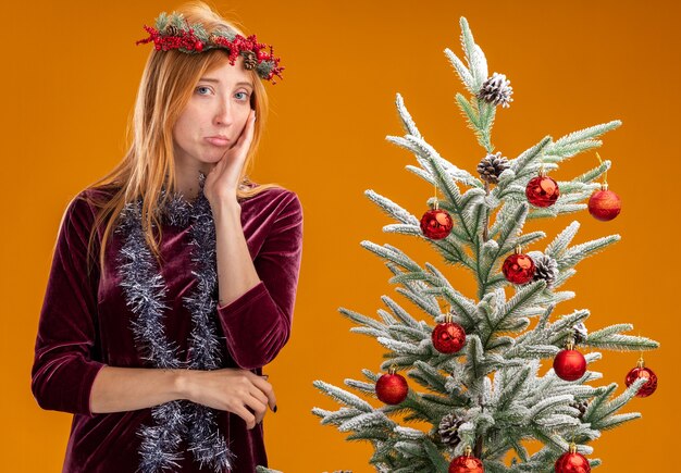 オレンジ色の壁に分離された頬に手を置き、赤いドレスと花輪を首につけた花輪を着てクリスマス ツリーの近くに立っている悲しい若い美しい女の子