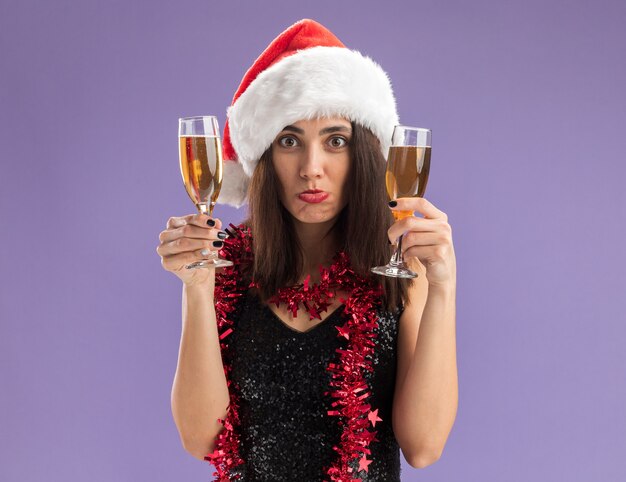 Sad young beautiful girl wearing christmas hat with garland on neck holding glass of champagne isolated on purple background