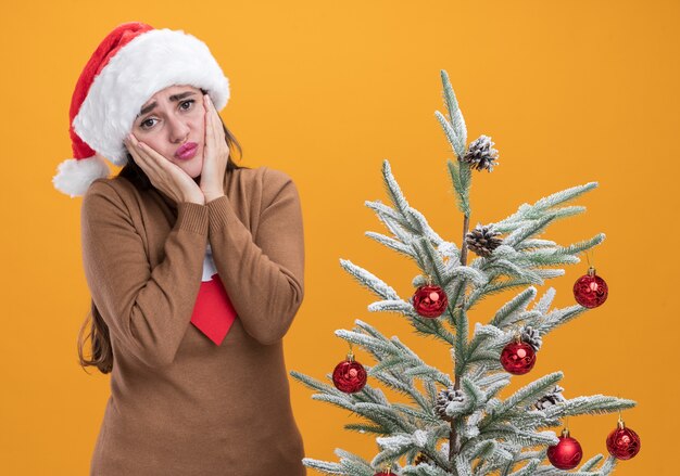 オレンジ色の背景で隔離の頬に手を置いてクリスマスツリーの近くに立っているネクタイとクリスマス帽子をかぶって悲しい若い美しい少女