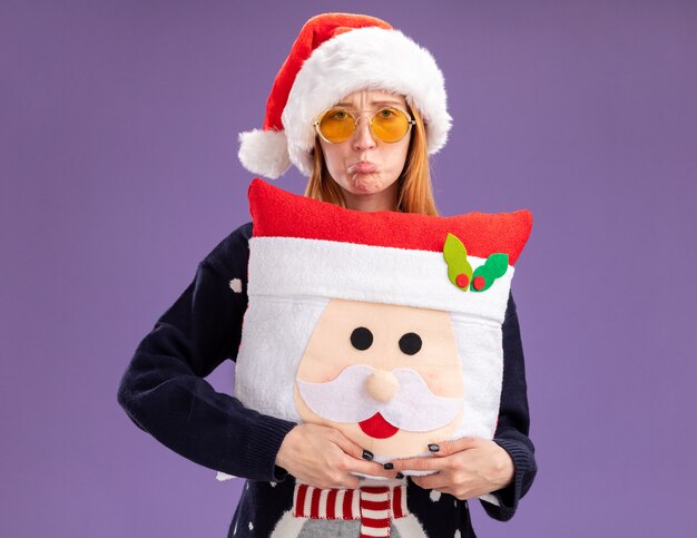 sad young beautiful girl wearing christmas sweater and hat with glasses holding christmas pillow isolated on purple wall