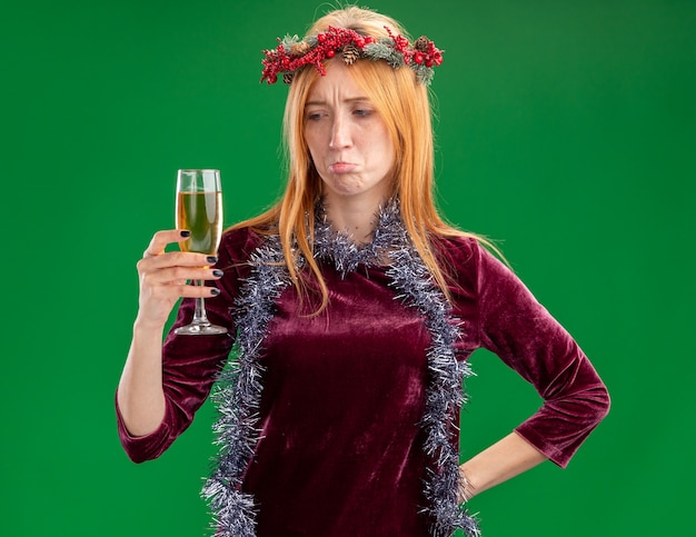 Sad young beautiful girl wearing red dress with wreath and garland on neck holding and looking at glass of champagne isolated on green background