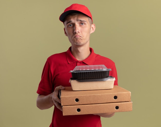 Sad young blonde delivery boy holds food packages on pizza boxes isolated on olive green wall with copy space