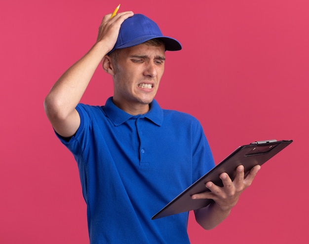 Sad young blonde delivery boy puts hand on head holding and looking at clipboard on pink 