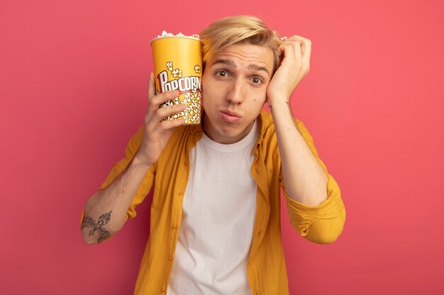Sad young blonde guy wearing yellow t-shirt holding bucket of popcorn around face isolated on pink