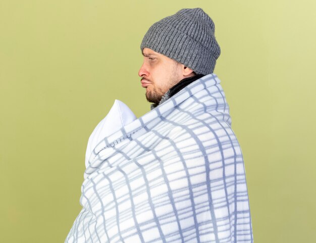 Sad young blonde ill man wearing winter hat and scarf wrapped in plaid stands sideways holding pillow isolated on olive green wall