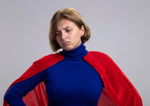 Sad young blonde superhero woman in red cape keeping hands on waist looking down isolated on white wall