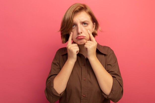 Sad young blonde woman looking at front pulling down eye lids isolated on pink wall with copy space