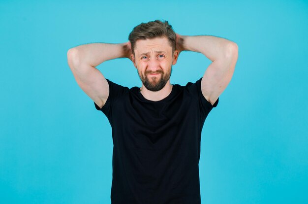 Sad young boy is holding hands behind head on blue background