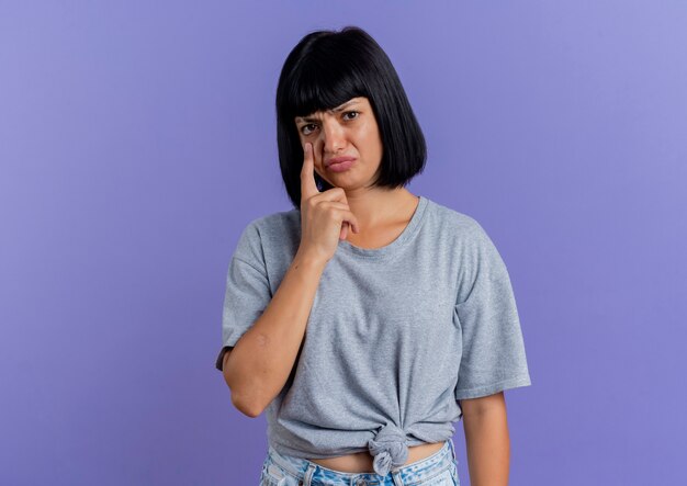 Sad young brunette caucasian girl puts finger on face isolated on purple background with copy space