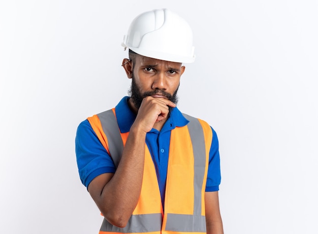 sad young builder man in uniform with safety helmet putting his hand on chin isolated on white wall with copy space
