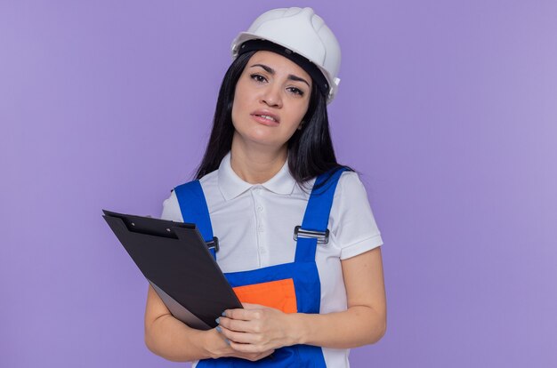 Sad young builder woman in construction uniform and safety helmet holding clipboard and pencil looking at front tired and bored standing over purple wall
