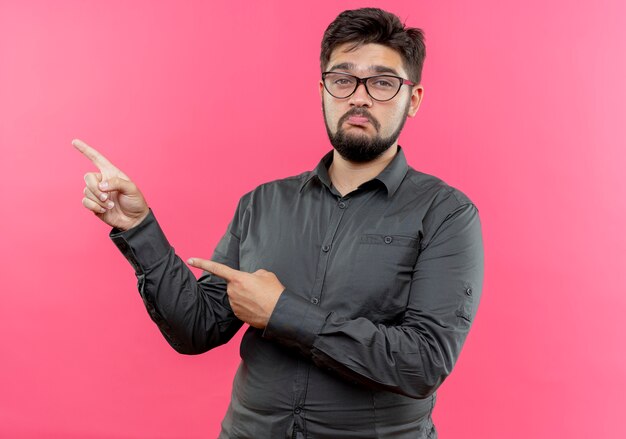 Sad young businessman wearing glasses points at side isolated on pink with copy space