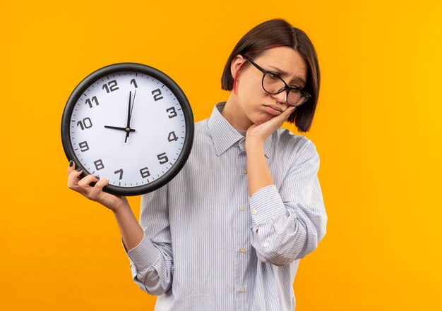 Sad young call center girl wearing glasses holding clock putting hand on cheek and looking down isolated on orange wall