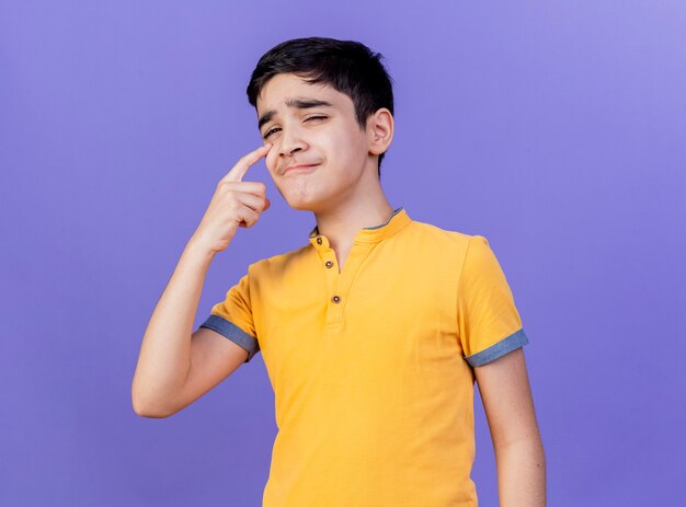 Sad young caucasian boy looking at camera putting finger under eye isolated on purple background